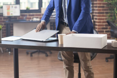 Young male employee with spectacles, carefully arranging his laptop at desk in brick wall workplace. Black businessman setting his digital device on table, preparing for work near office documents.