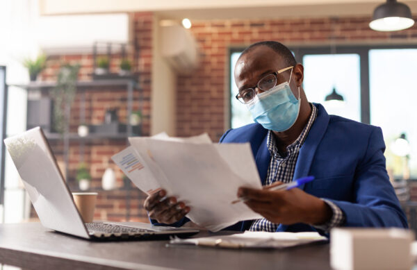 African american man with face mask analyzing documents to plan marketing strategy. Company employee working with laptop and doing paperwork for project during coronavirus pandemic.