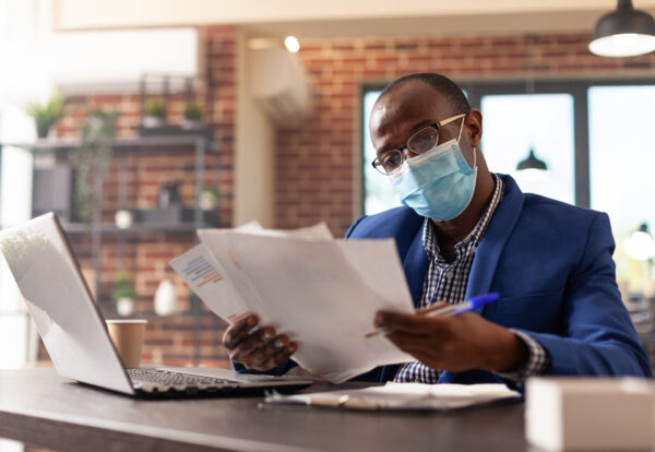 African american man with face mask analyzing documents to plan marketing strategy. Company employee working with laptop and doing paperwork for project during coronavirus pandemic.