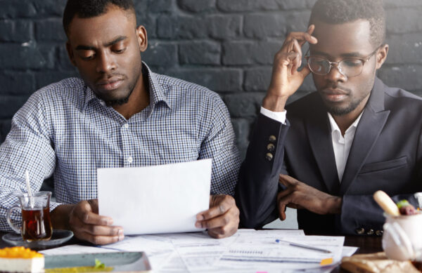 People, business and partnership concept. African-American manager wearing glasses and his colleague having tired and stressed looks while reviewing finances, working through papers together