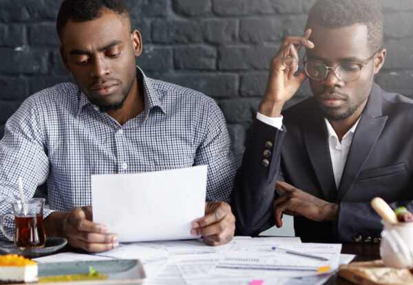 People, business and partnership concept. African-American manager wearing glasses and his colleague having tired and stressed looks while reviewing finances, working through papers together