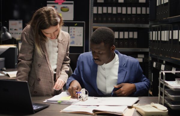 In evidence room, multiethnic private detectives work together to perform criminal investigations. African american and caucasian inspectors use laptop and case files to gather clues and information.